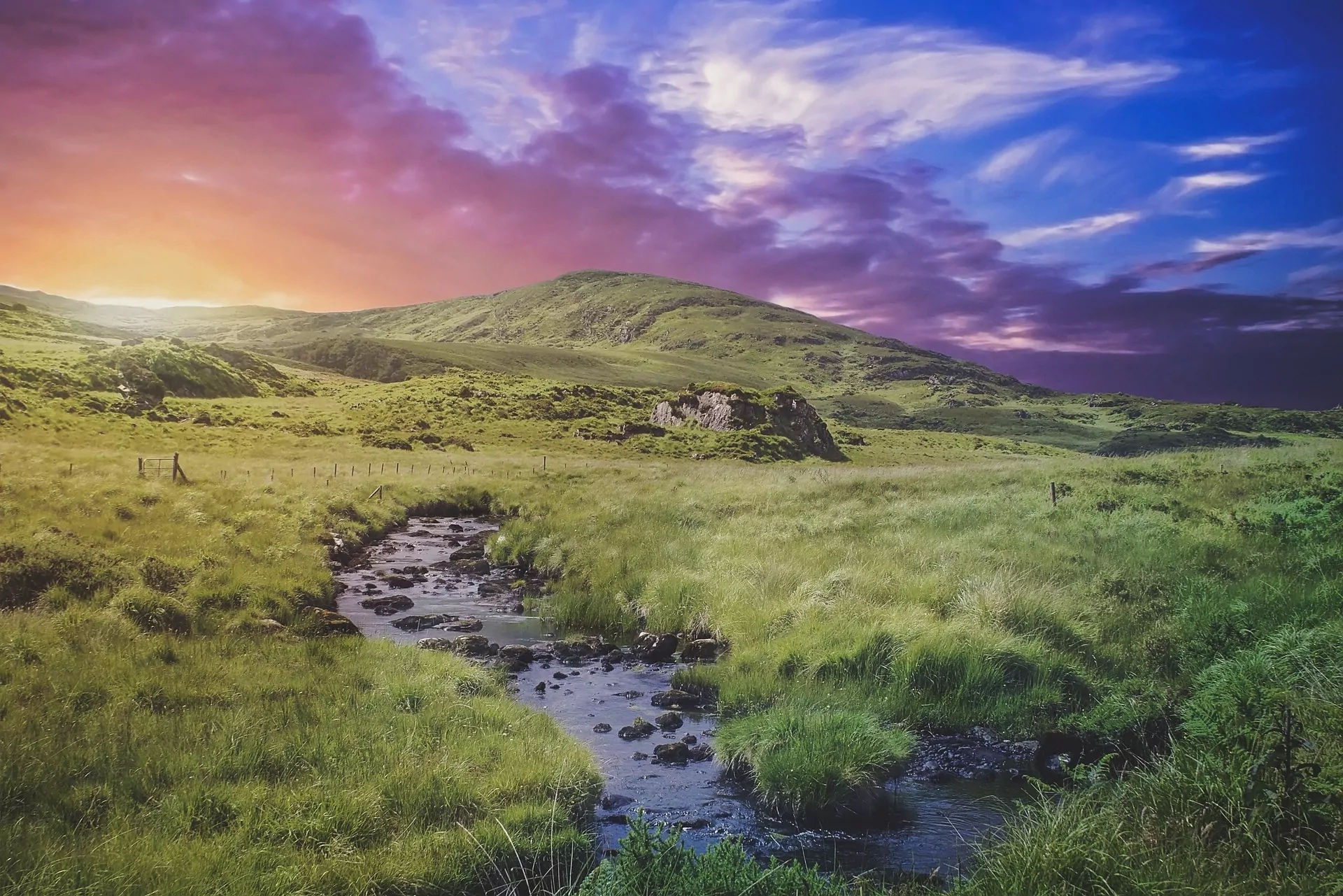 An image of a sun setting over the English countryside. The rich array of colours marbled across the evening sky is beautiful. A stream cuts down the centre of the frame making a calm and tranquil scene. David Attenborough series: Wild Isles.