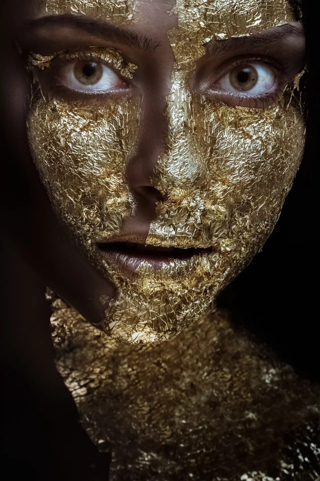 A close-up image of a woman's face covered in gold leaf. She looks directly into the camera. Time-Lapse of costume and make-up transformations.