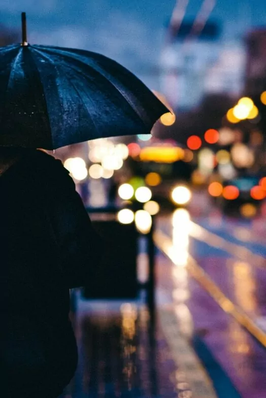 An image of a pedestrian standing under an umbrella at nighttime. The glare from street lights and buildings are reflected in puddles. A beautiful urban image at night. BBC Panorama's 15 Minute City.