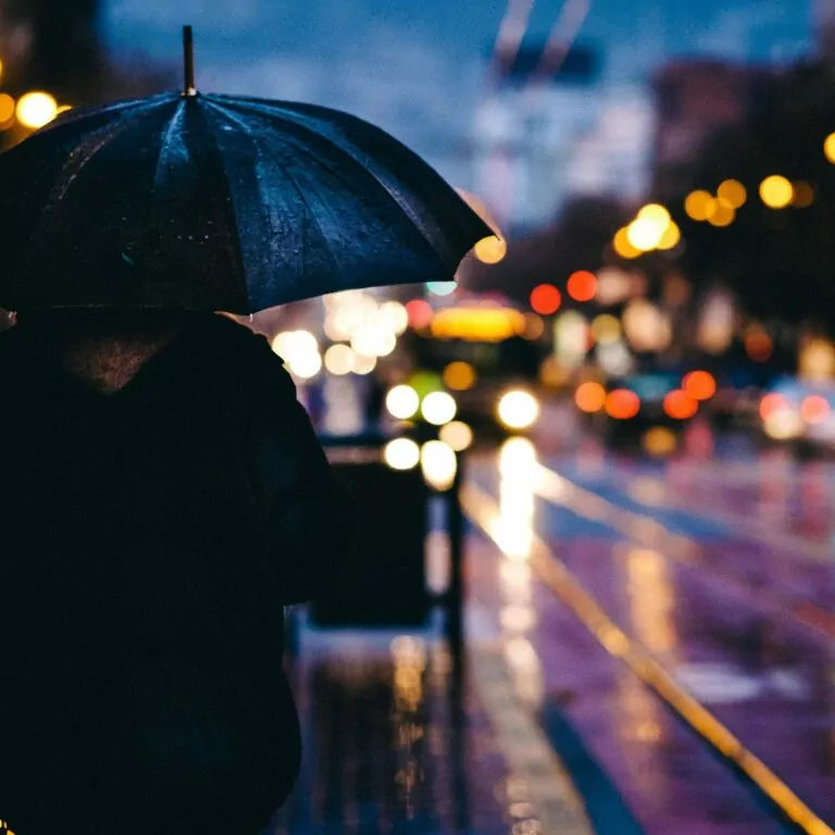 An image of a pedestrian standing under an umbrella at nighttime. The glare from street lights and buildings are reflected in puddles. A beautiful urban image at night. BBC Panorama's 15 Minute City.