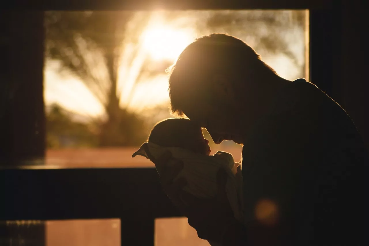 An image of a Father holding a newborn baby. Sunlight shines from directly behind them casting the figures in shadow. The Father rests his forehead on the baby in their embrace.