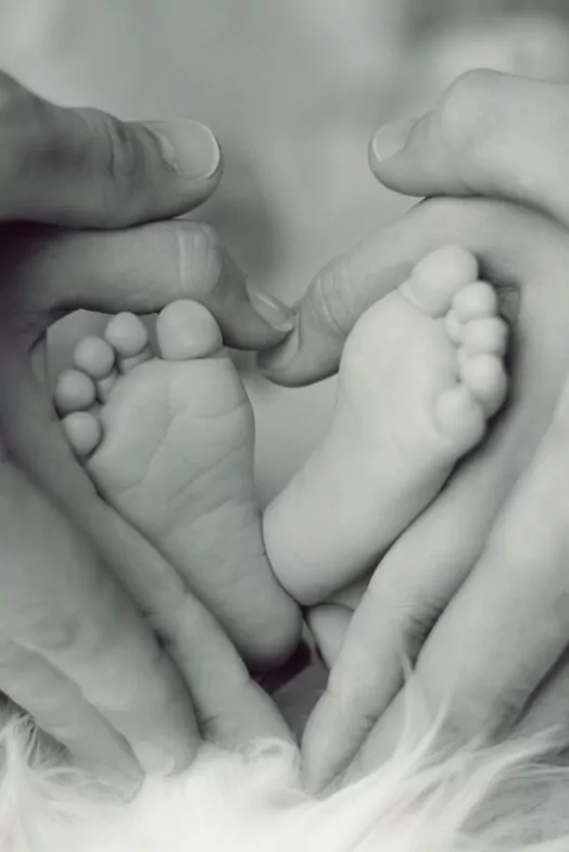 An image of a newborn baby's feet held my it's Mother's and Father's hands. The feet and hands entwined make a heart shape.