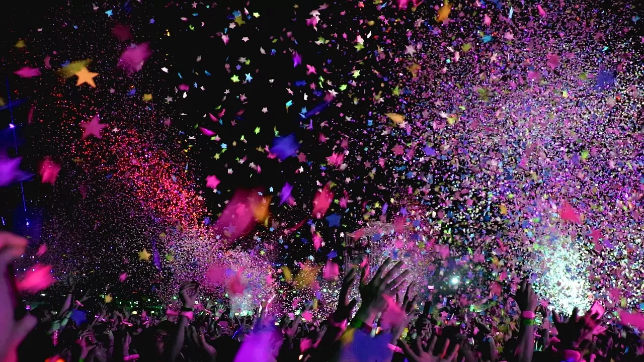 An image of festival-goers dancing in a crowd. Bright pink confetti billows down over head making it a jubilant scene.