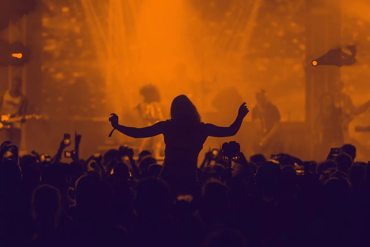Orange hued stage lights illuminate a festival-goer who is lifted onto the shoulders of a friend. 