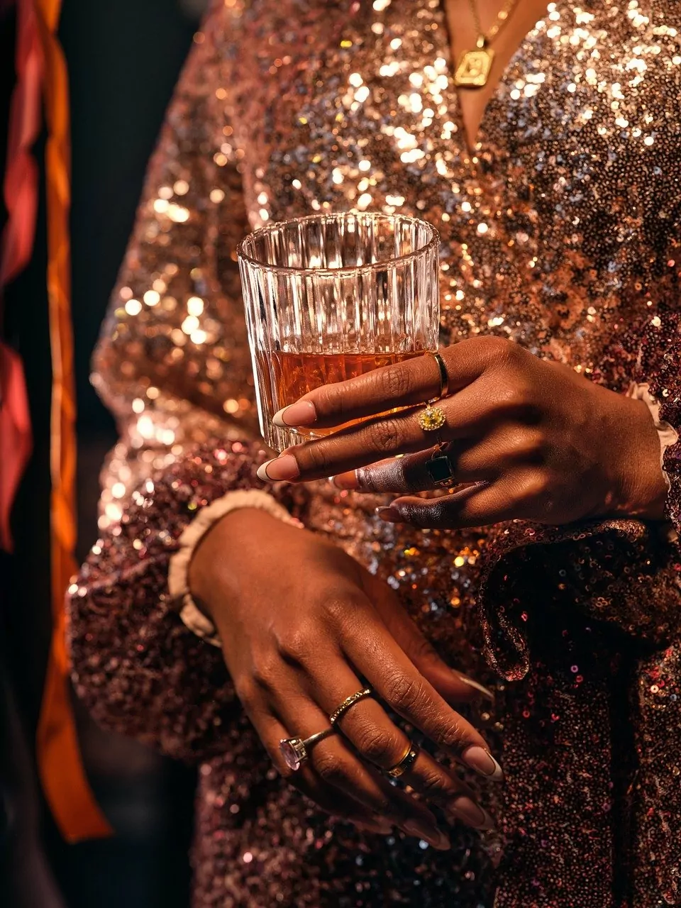 A woman in glamorous sequinned attire ta New Year's Eve celebration. The woman holds a glass of whiskey and her dazzling jewellery is in shot.